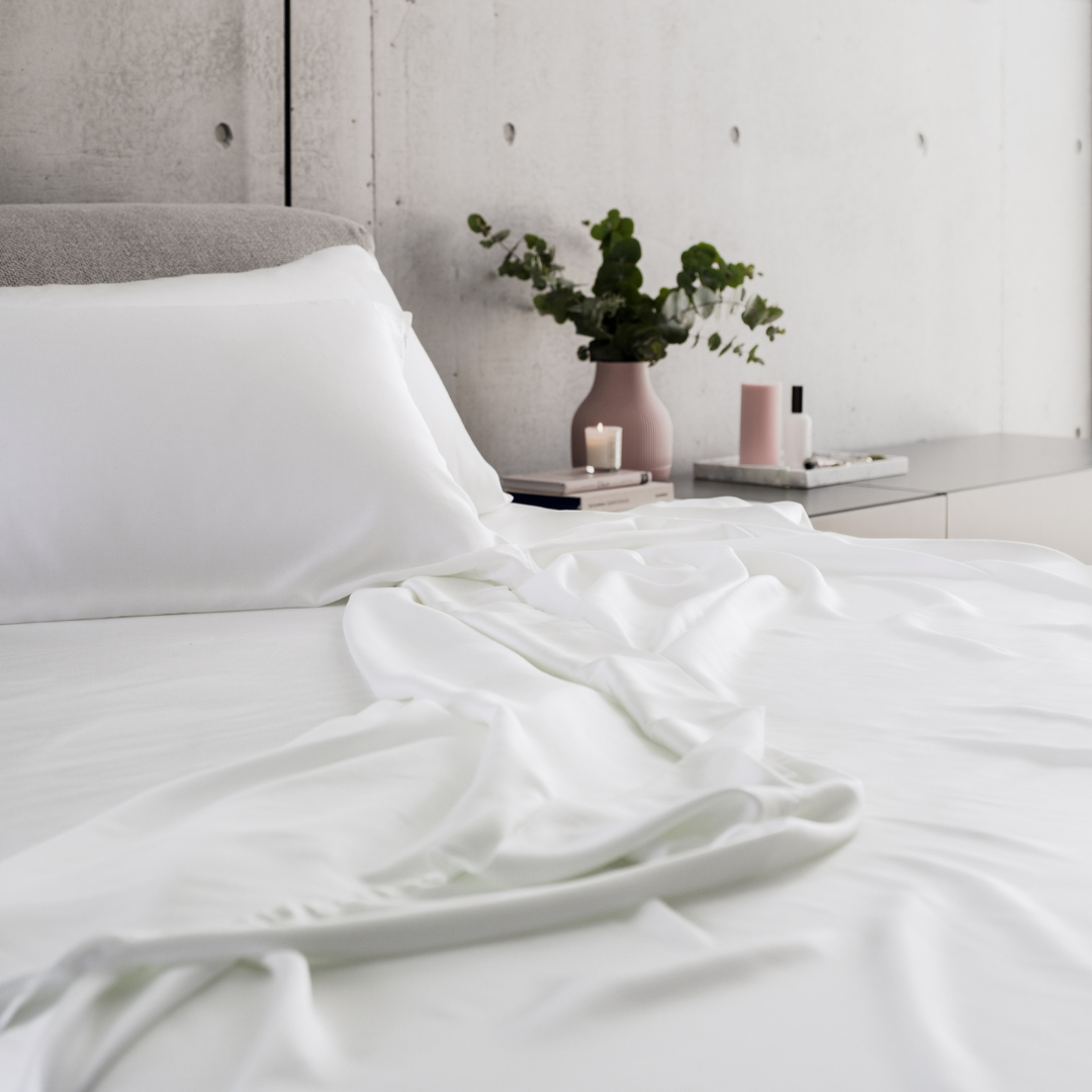 A neatly made bed with white sheets and pillows sits in a modern bedroom. On the bedside table are a small pink vase with green foliage, a lit candle, books, and a pink cylindrical container.